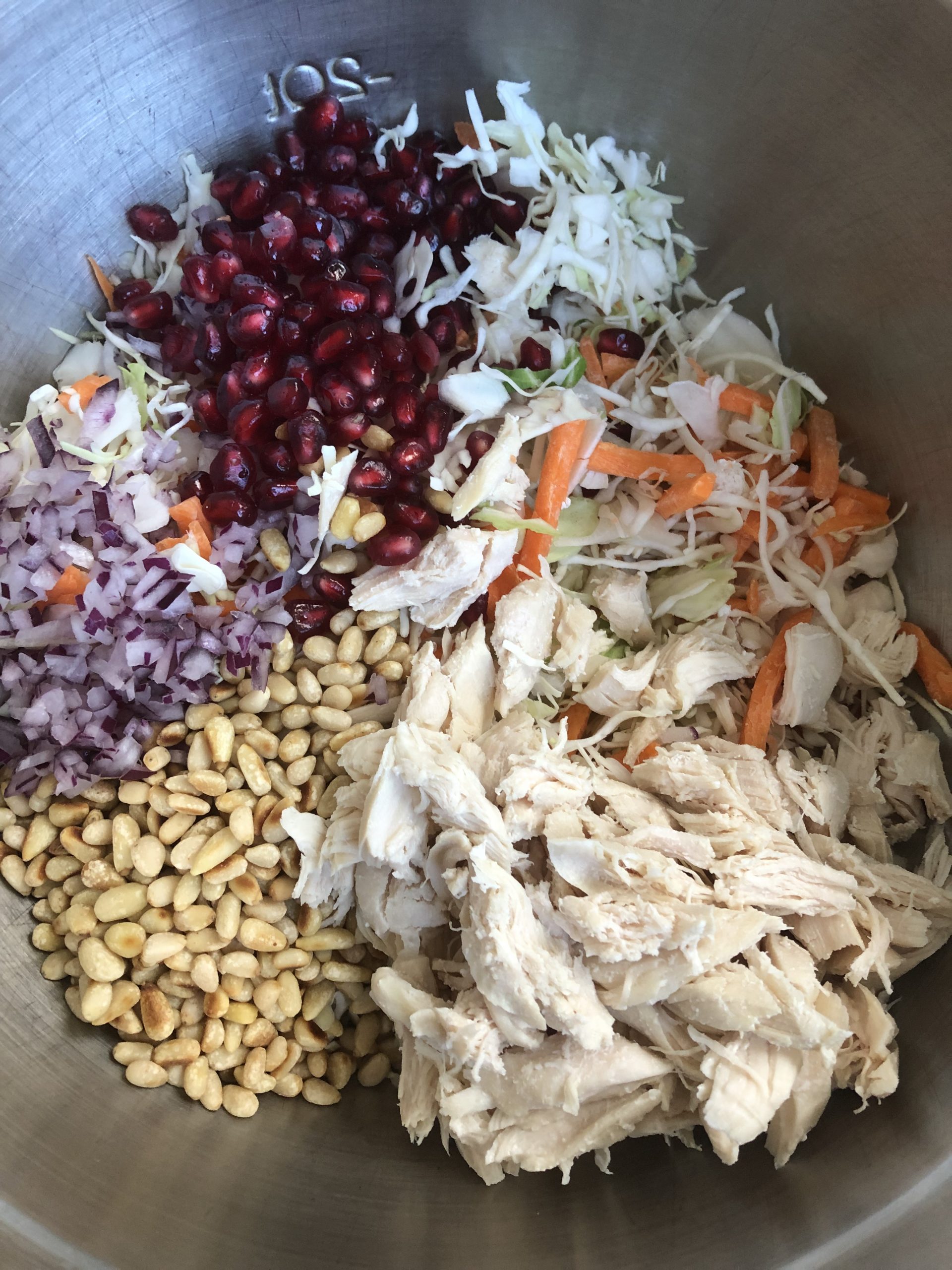 A large bowl with shredded chicken, cole slaw mix, pomegranate, red onion, and pine nuts.