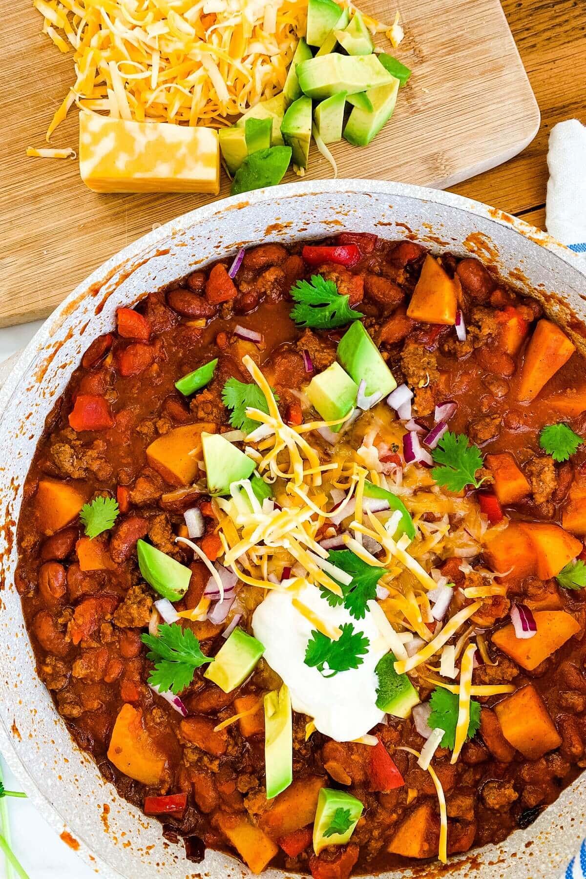 Beef and butternut squash chili in a pot topped with avocado, cheese and sour cream.