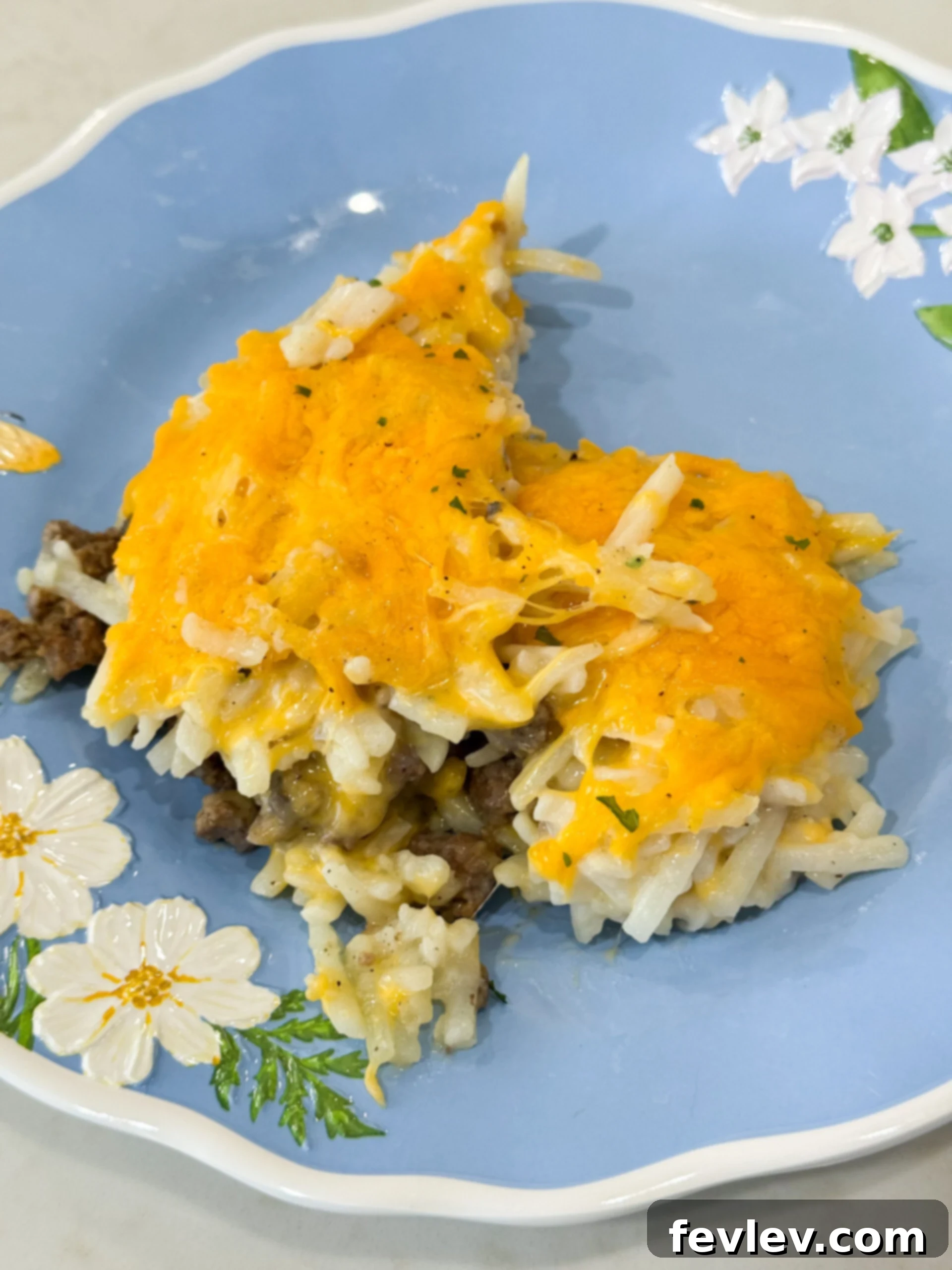 Golden Hashbrown Beef Bake 2 A delicious, baked Hashbrown Hamburger Casserole served on a white plate, topped with golden melted cheddar cheese and fresh parsley garnish. Steam rises gently from the warm, hearty meal.