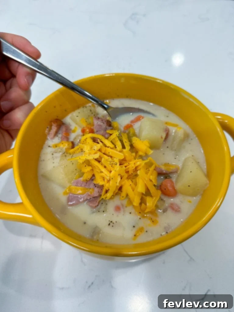 A steaming bowl of creamy Ham and Potato Soup, topped with melted shredded cheese and fresh green herbs.
