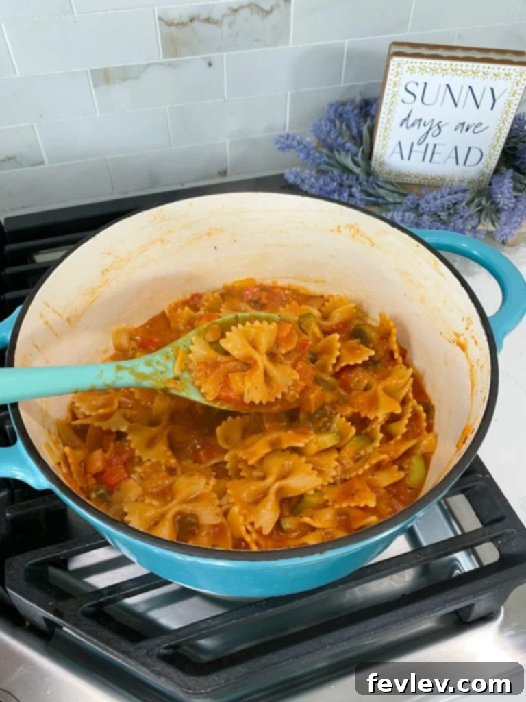 Assortment of fresh ingredients for Italian vegetable pasta, including bell peppers, zucchini, mushrooms, onions, garlic, tomato paste, and uncooked bow-tie pasta, laid out on a kitchen counter.