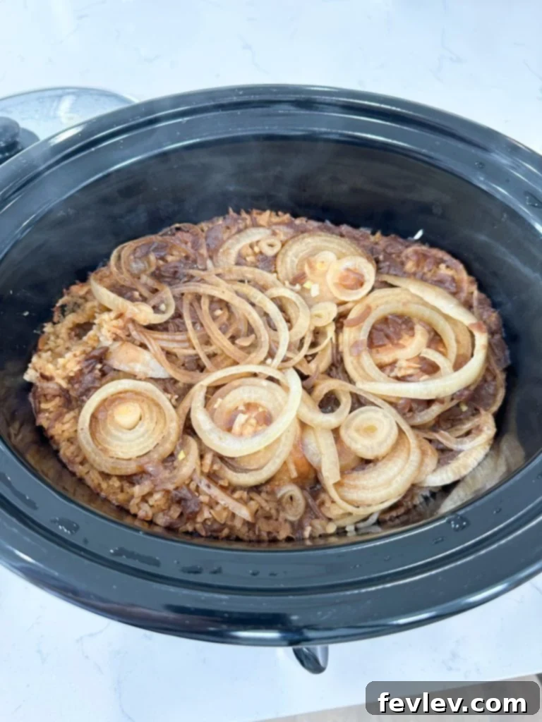 Slow cooker bowl filled with layered raw chicken, sliced onions, rice, onion soup mix, beef broth, and canned French onion soup, before cooking begins.