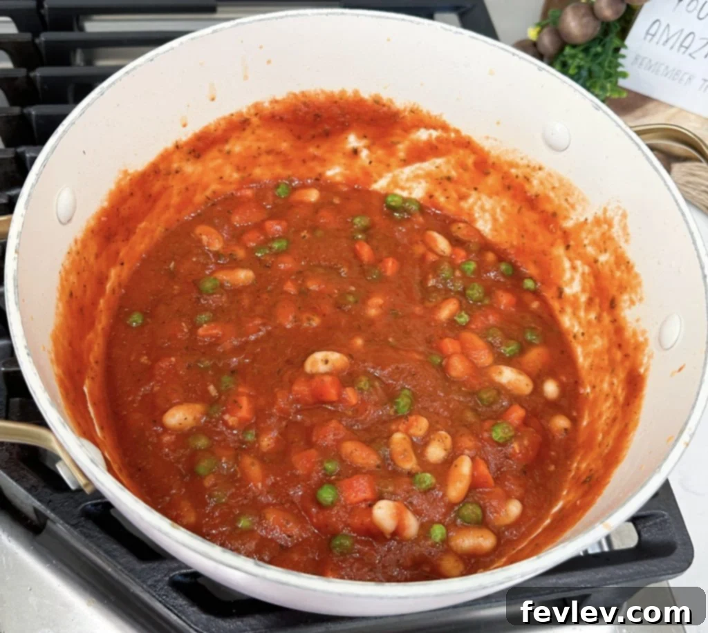 A pot on the stove filled with rich red pasta sauce, white beans, and mixed frozen vegetables simmering together.