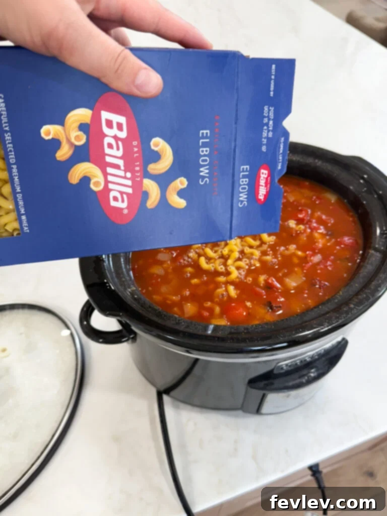Uncooked elbow macaroni being stirred into the simmering chili in a slow cooker.