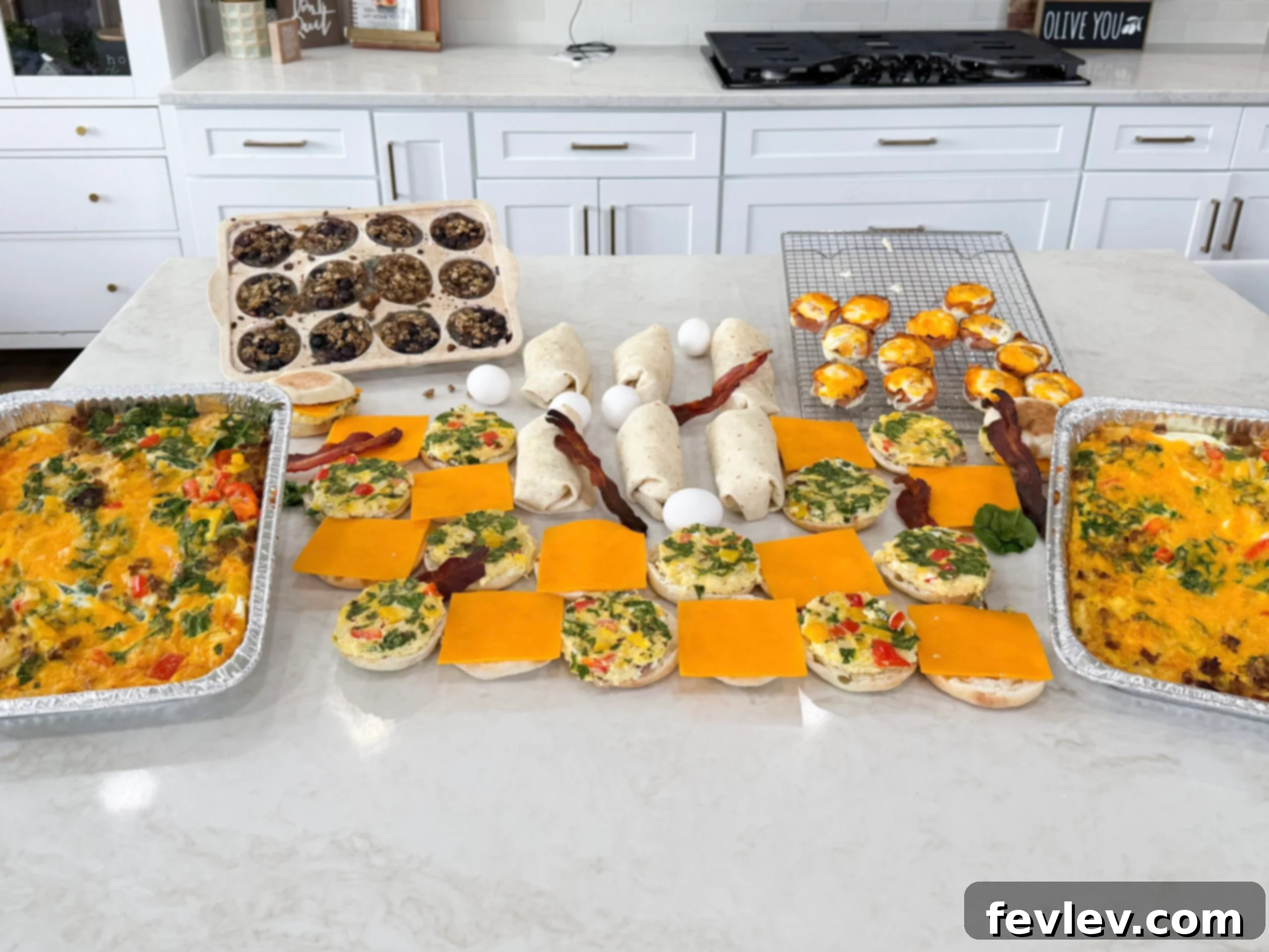 Effortless Breakfast Meal Prep 2 A vibrant collection of completed breakfast meal prep items, including two savory breakfast casseroles, neatly arranged baked blueberry oatmeal cups in a muffin tin, golden ham and egg bites cooling on a rack, freshly wrapped breakfast burritos, and stacked breakfast sandwiches, all presented on a clean white countertop.