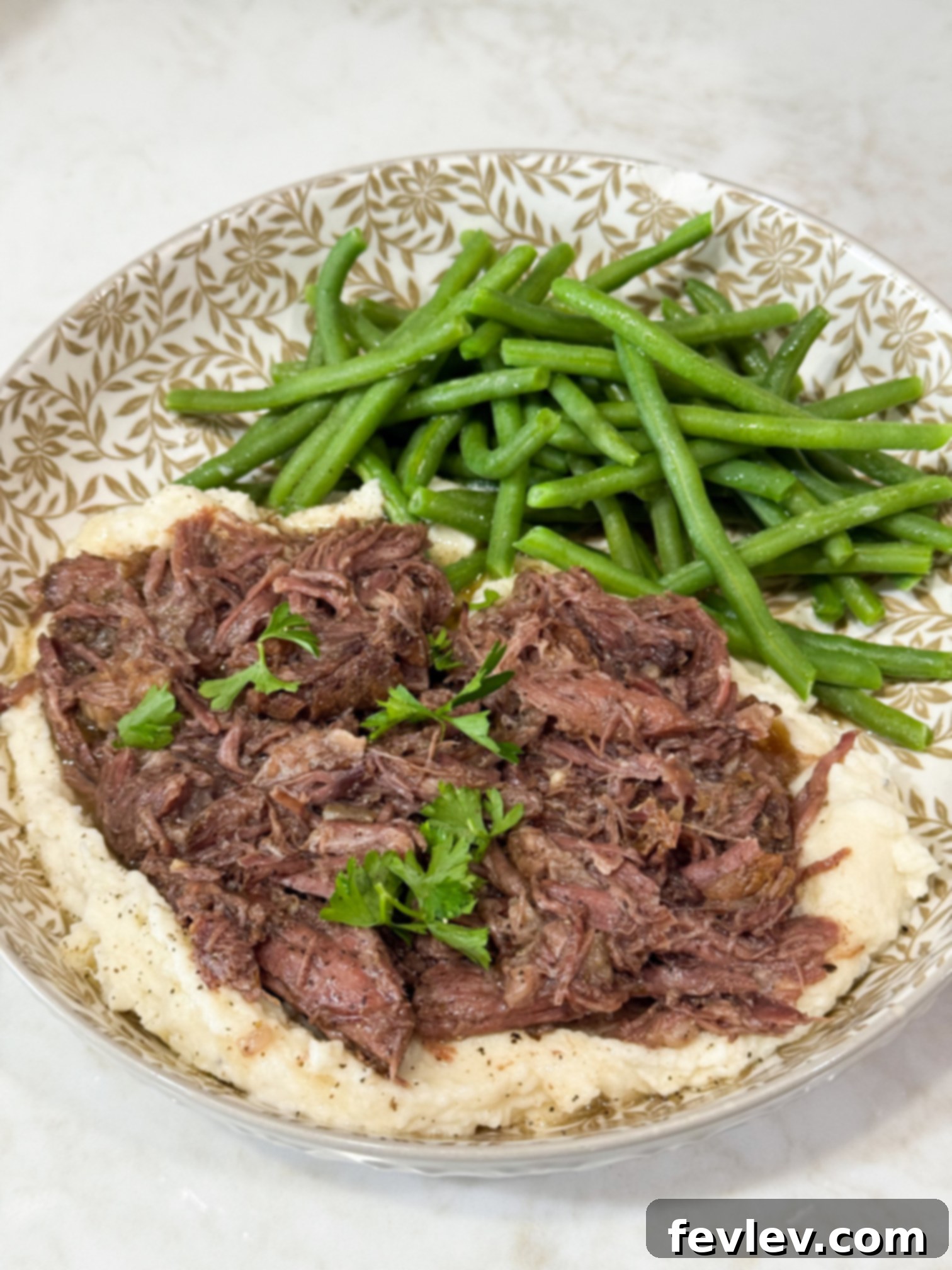 Tender shredded beef served over mashed potatoes with green beans, presented on a stylish grey and white plate. Perfect for a hearty meal.