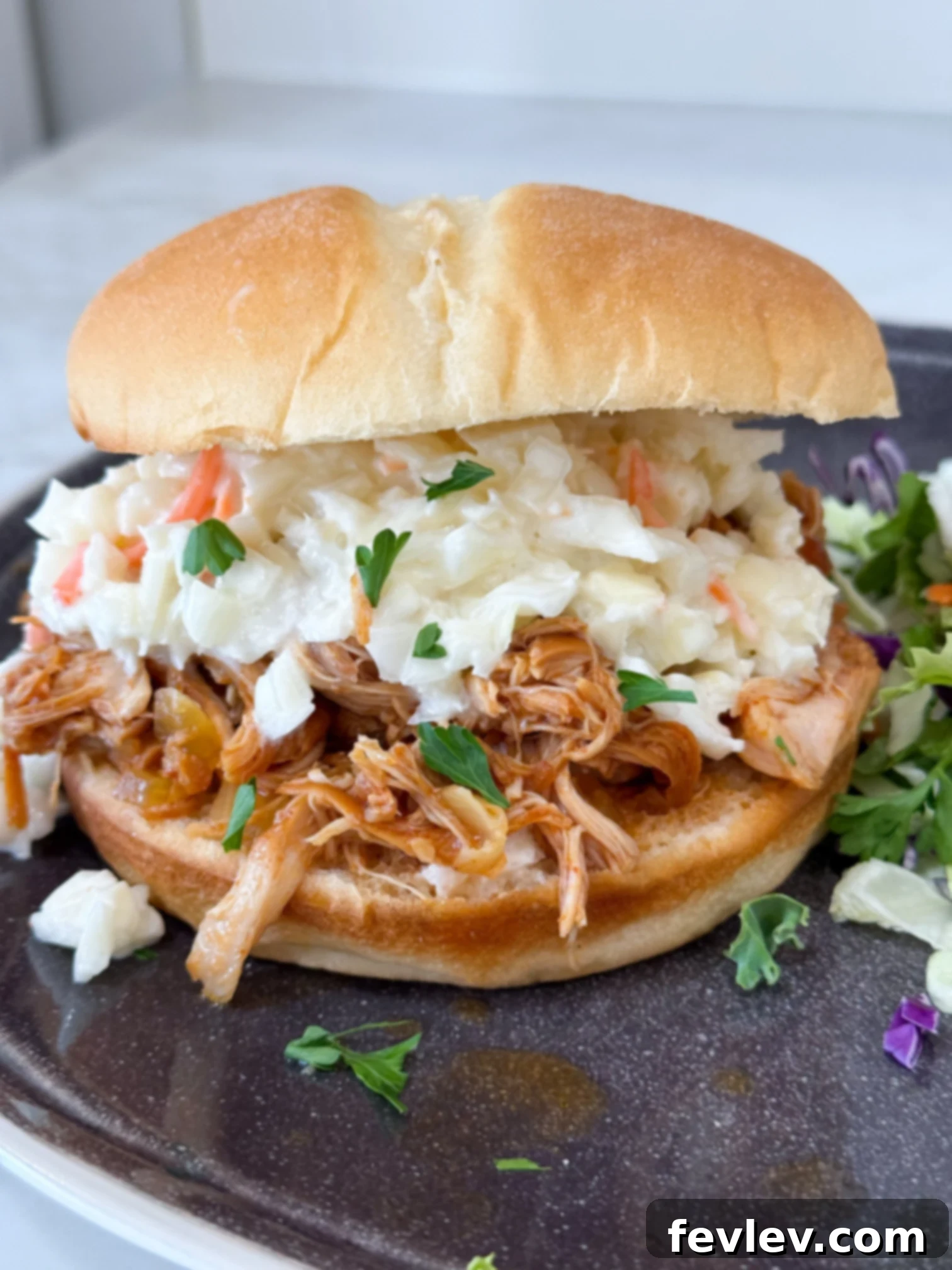 Close-up of Slow Cooker Hawaiian Barbecue Chicken Sandwiches served on a black plate with a fresh green salad on a grey kitchen counter, highlighting the vibrant colors and appealing texture of the shredded chicken on a bun.