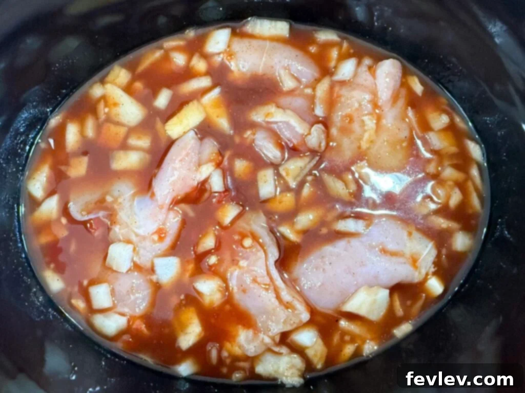 Raw chicken thighs coated in Hawaiian sauce and diced onions at the bottom of a slow cooker, ready for cooking.