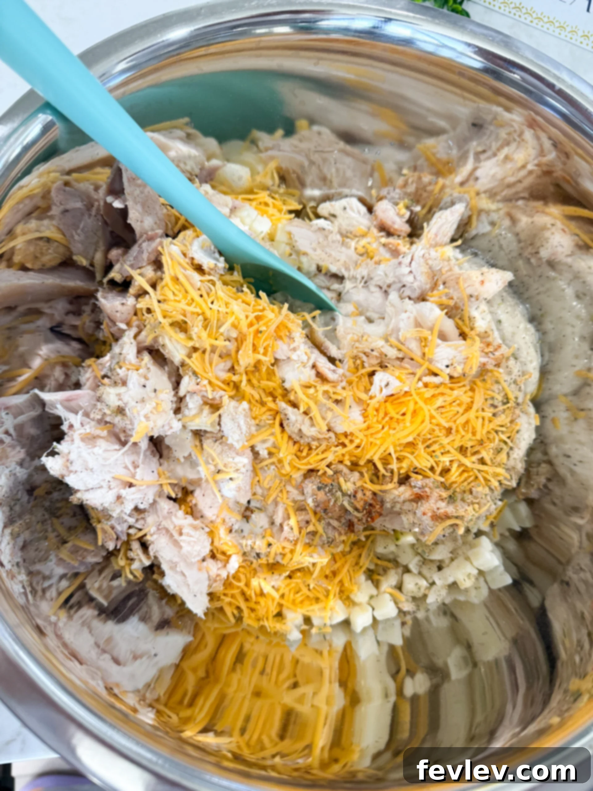 A large mixing bowl containing shredded chicken, frozen diced hash browns, cream of mushroom soup, French onion dip, cheddar cheese, and various seasonings, all ready to be combined.