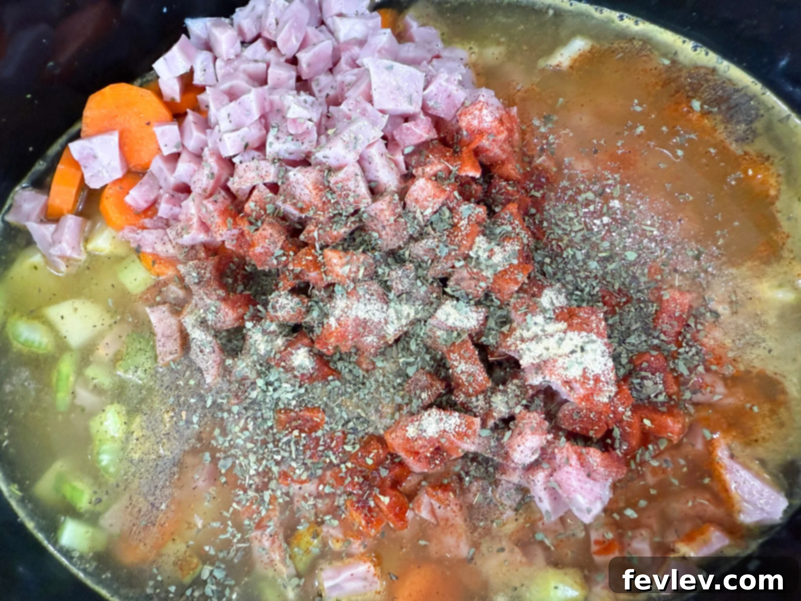 Chopped potatoes, onions, carrots, celery, and ham layered at the bottom of a slow cooker with chicken broth, ready for cooking