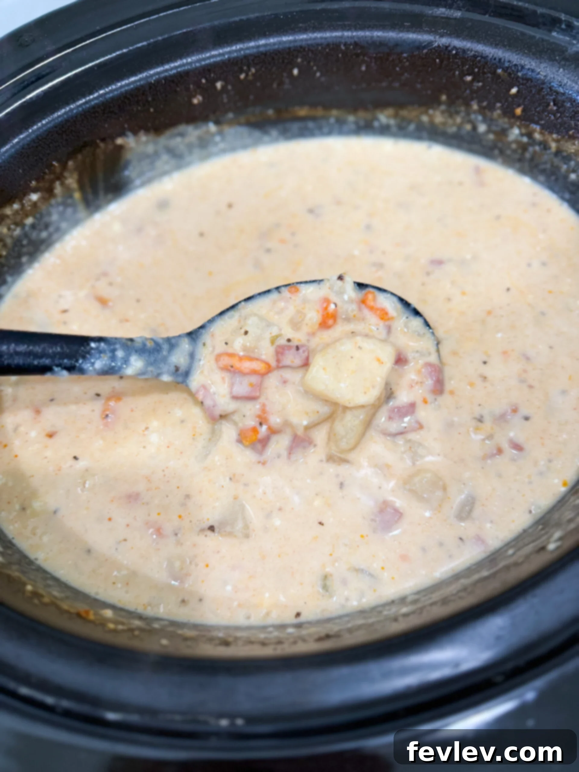 A cook stirring shredded cheddar cheese and sour cream into the bubbling ham and potato soup in a slow cooker, creating a rich and creamy texture