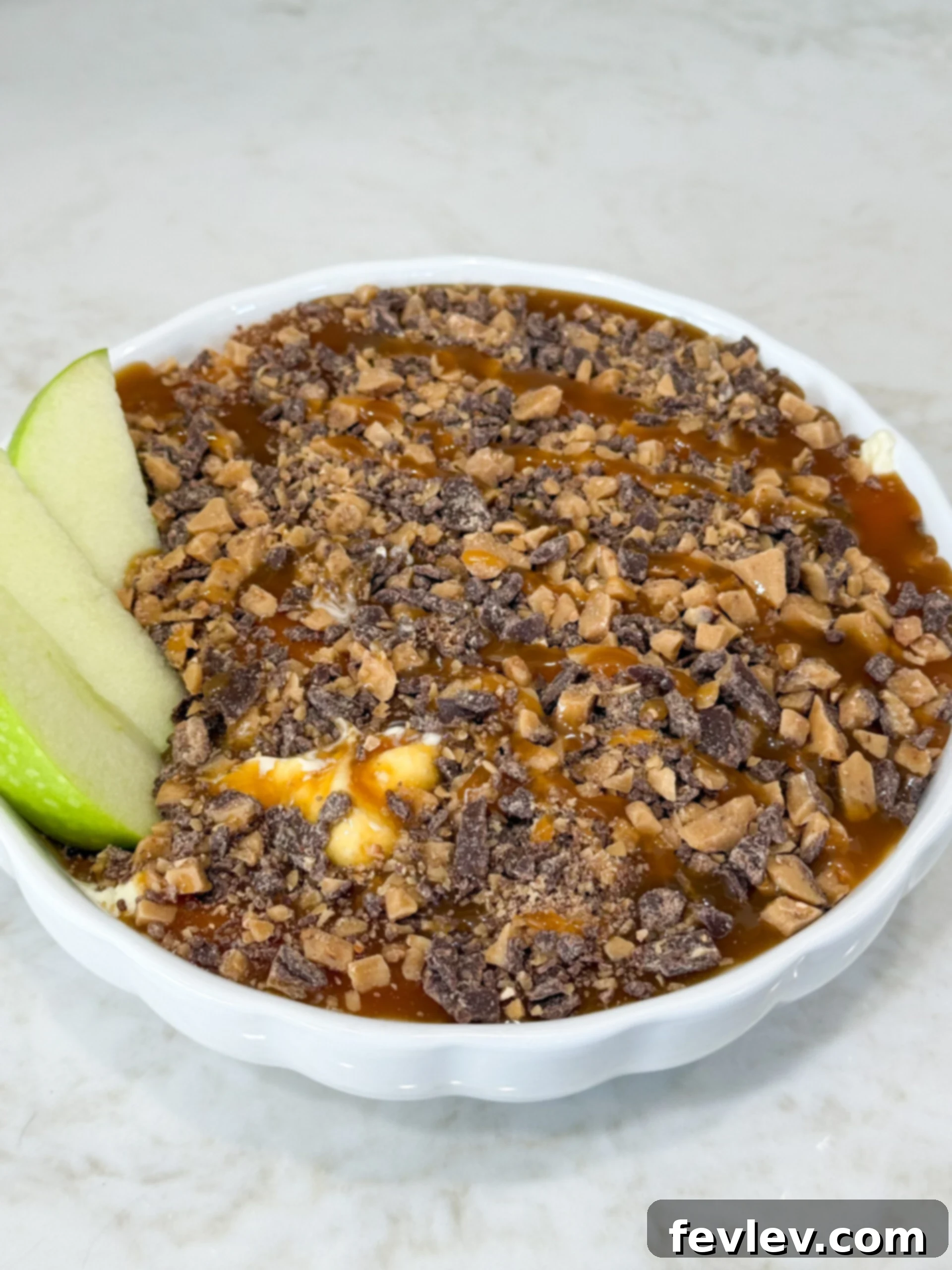 Close-up of a creamy Caramel Heath cheesecake dessert dip, generously topped with crunchy Heath toffee bits and caramel drizzle, served alongside fresh apple slices for dipping.