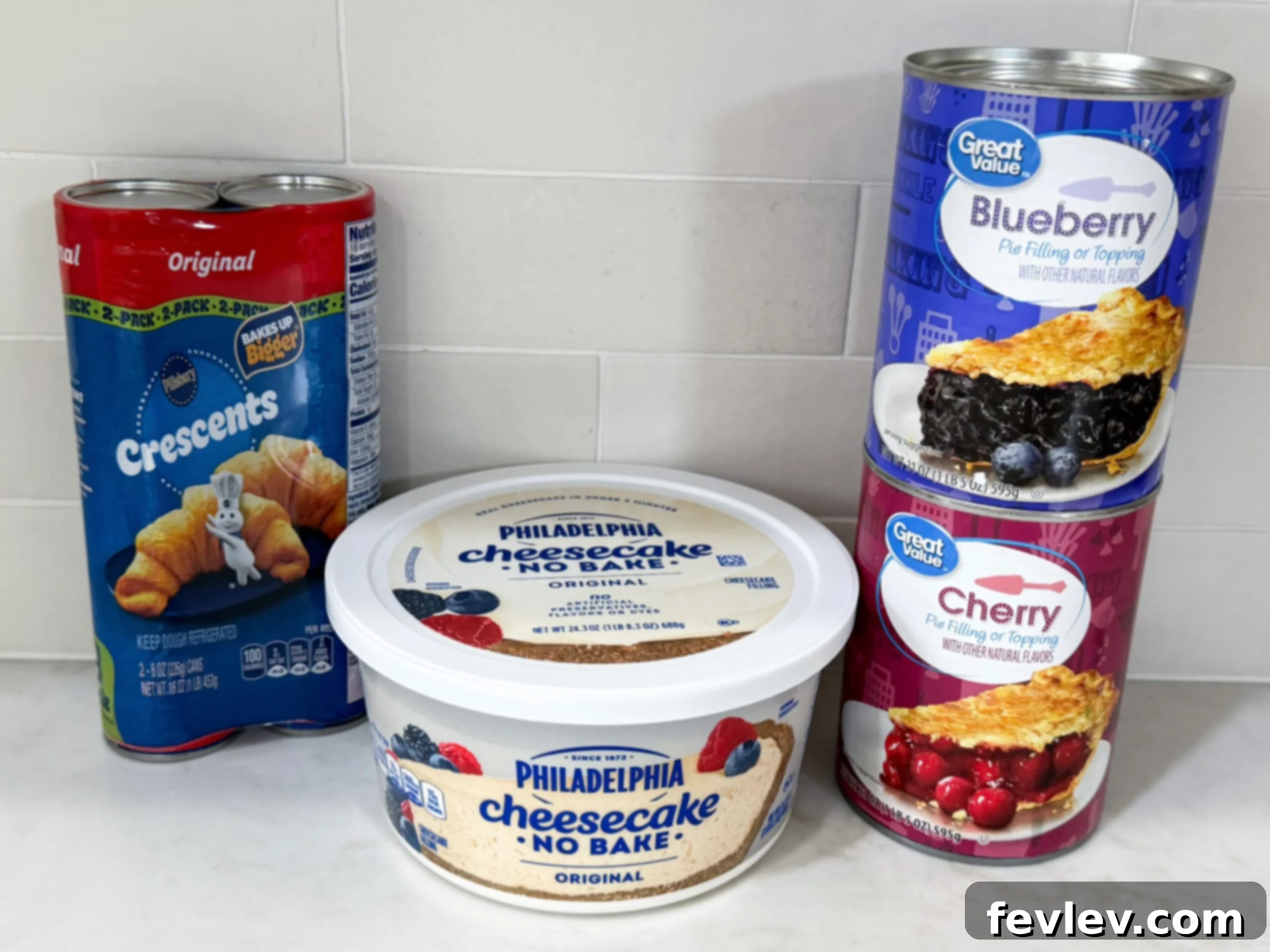 All the simple ingredients for mini cheesecake pies laid out on a kitchen counter: crescent dough, cheesecake filling, fruit topping, and powdered sugar.