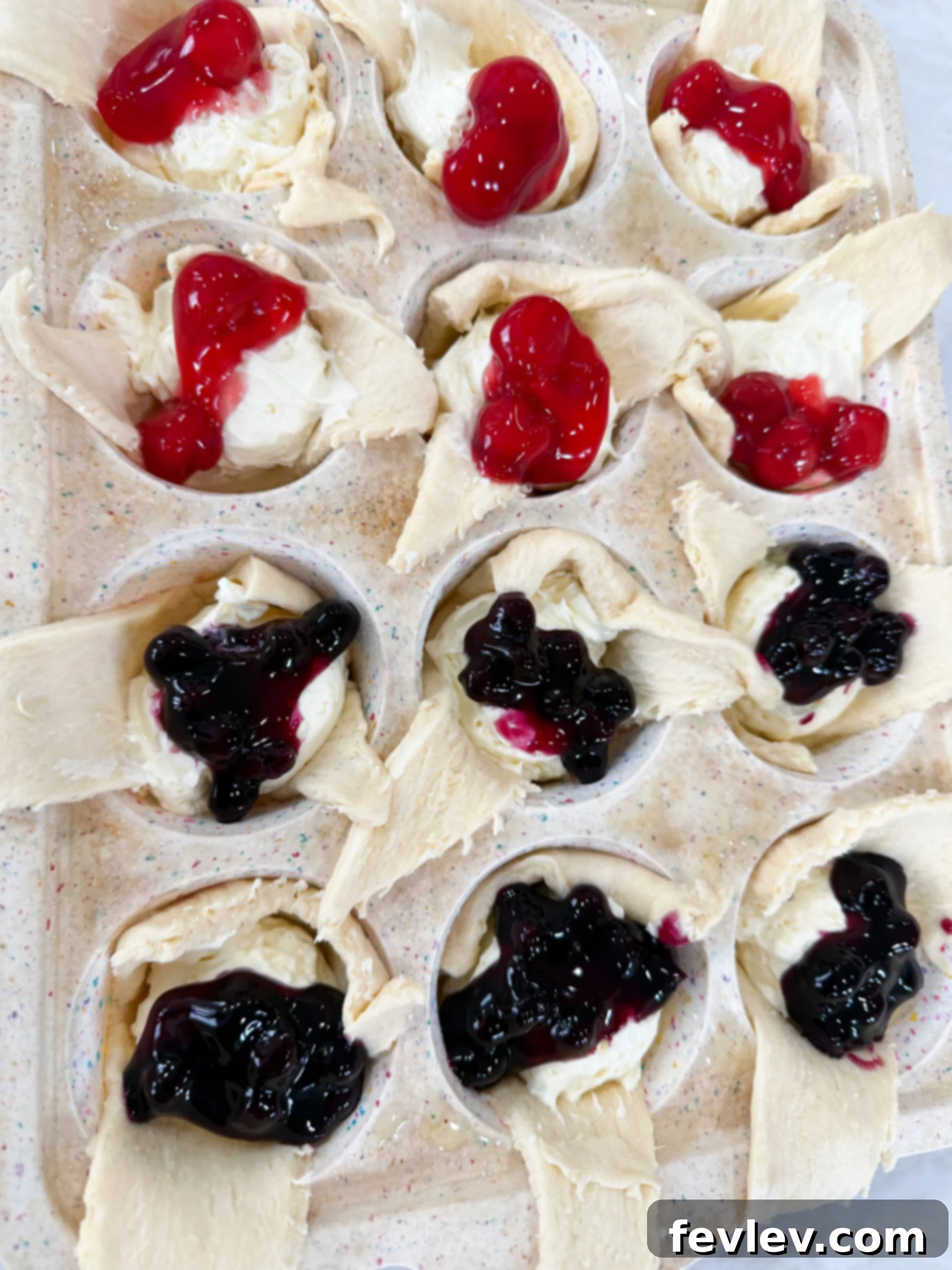 Vibrant blueberry and raspberry fruit pie filling being carefully placed on top of the cheesecake layer in crescent dough cups.