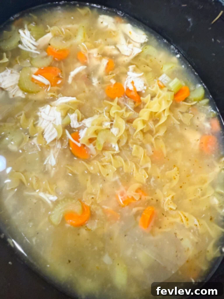 Shredded chicken and egg noodles being stirred into the hot, flavorful chicken noodle soup in the slow cooker.