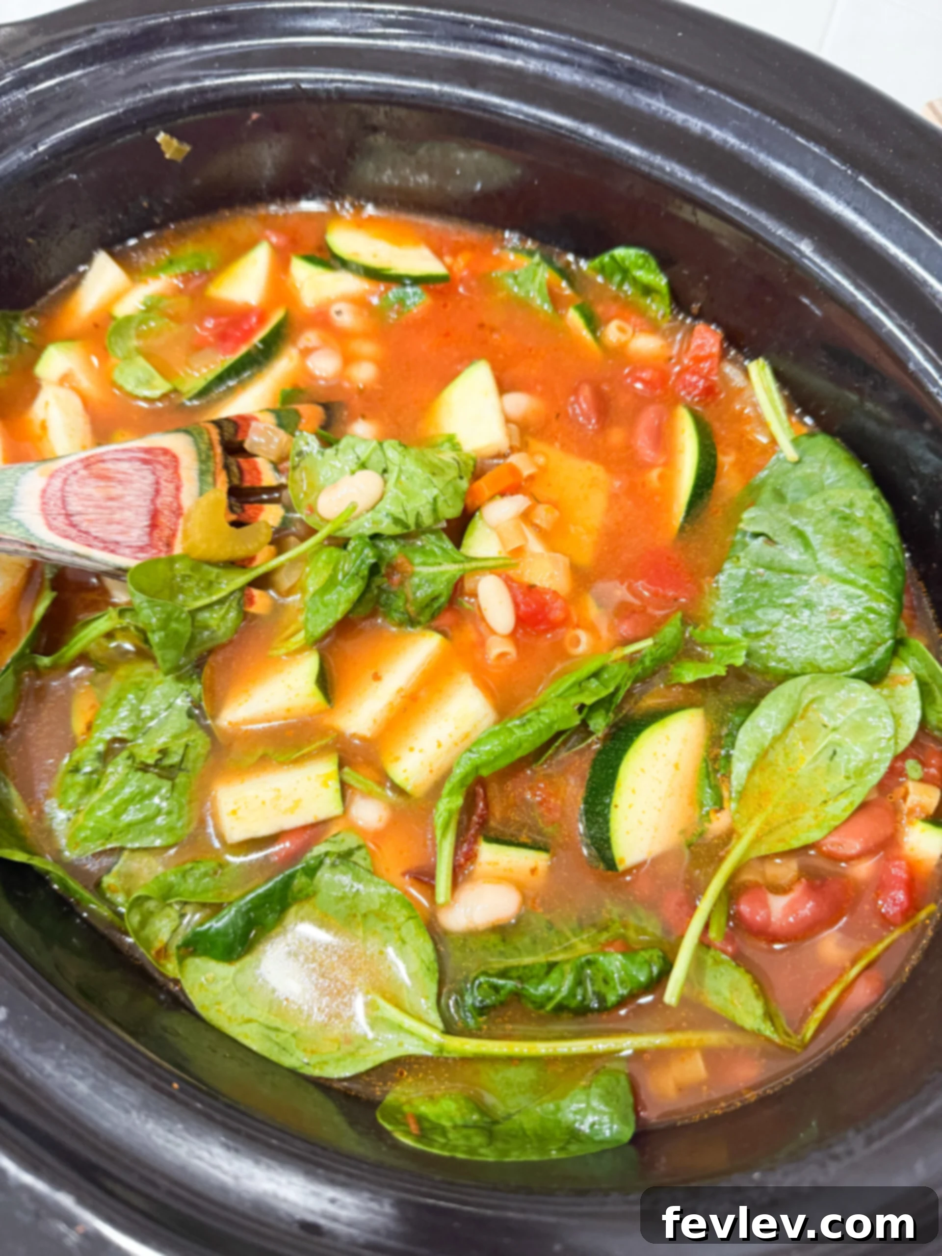The slow cooker filled with minestrone ingredients, cooking until vegetables are tender, followed by stirring in beans, zucchini, pasta, and spinach.