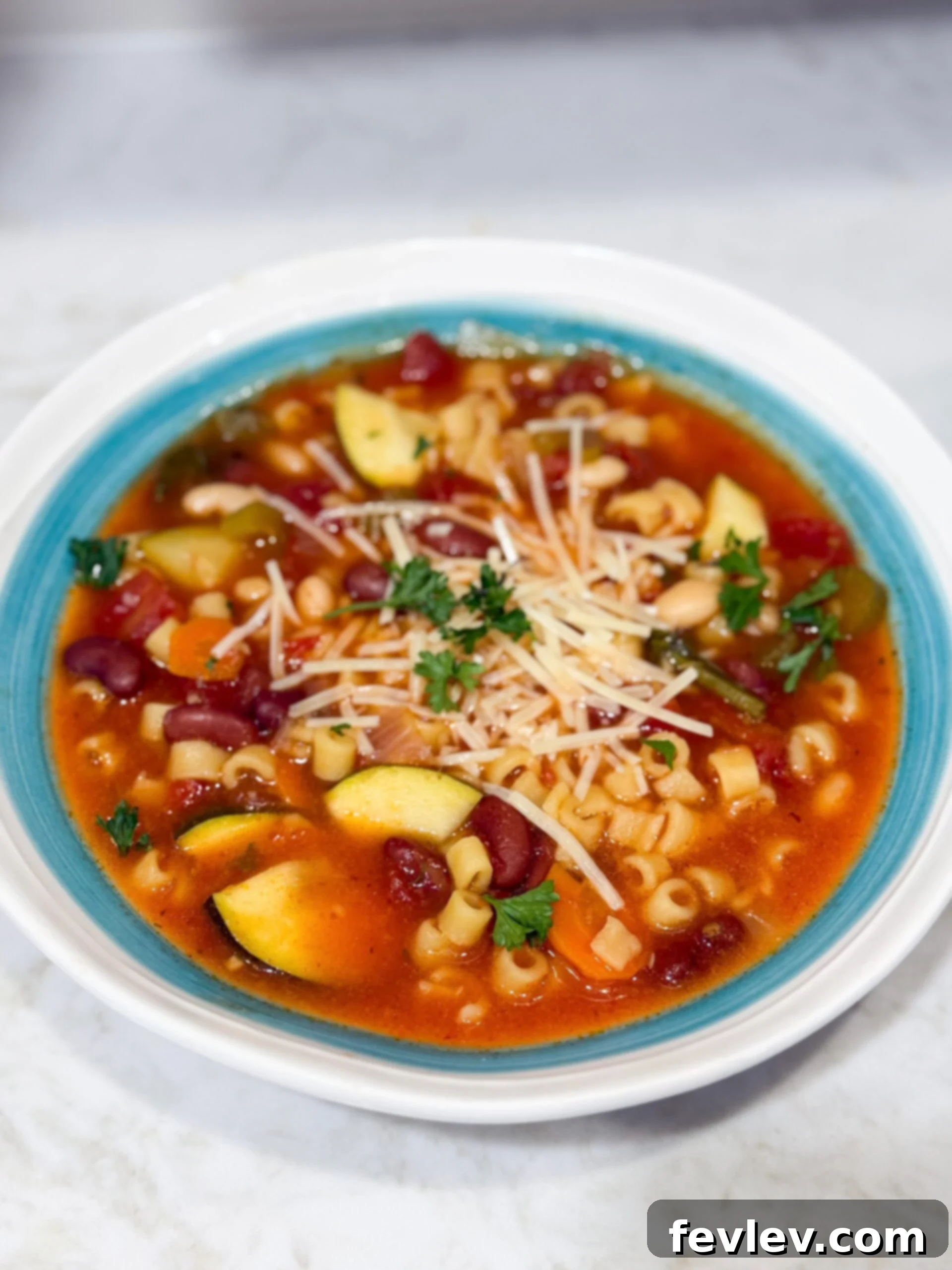 A serving of Slow Cooker Minestrone Soup in a white bowl, topped with a generous sprinkle of fresh Parmesan cheese and a sprig of rosemary.