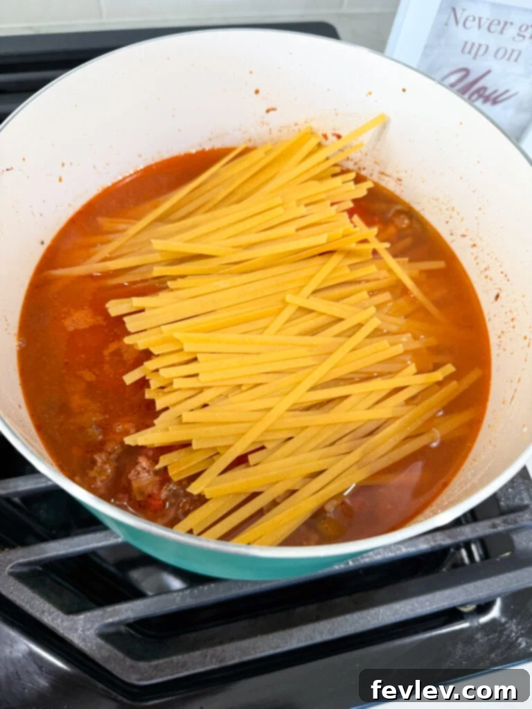 Diced tomatoes, Italian seasoning, water, and broken fettuccine noodles added to the browned beef and vegetable mixture in a pot, ready to simmer.